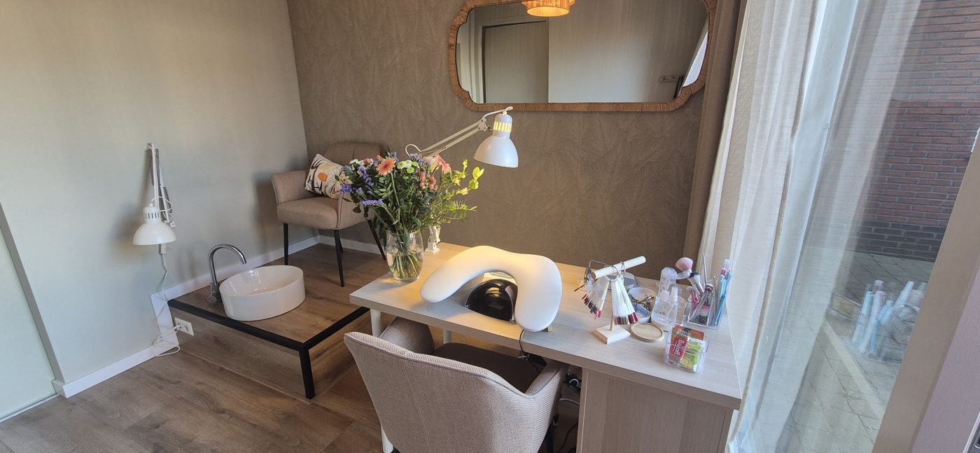 De salon: een witte werktafel met nagellamp en bloemen, een stoel, een spiegel met gouden rand en een groot raam